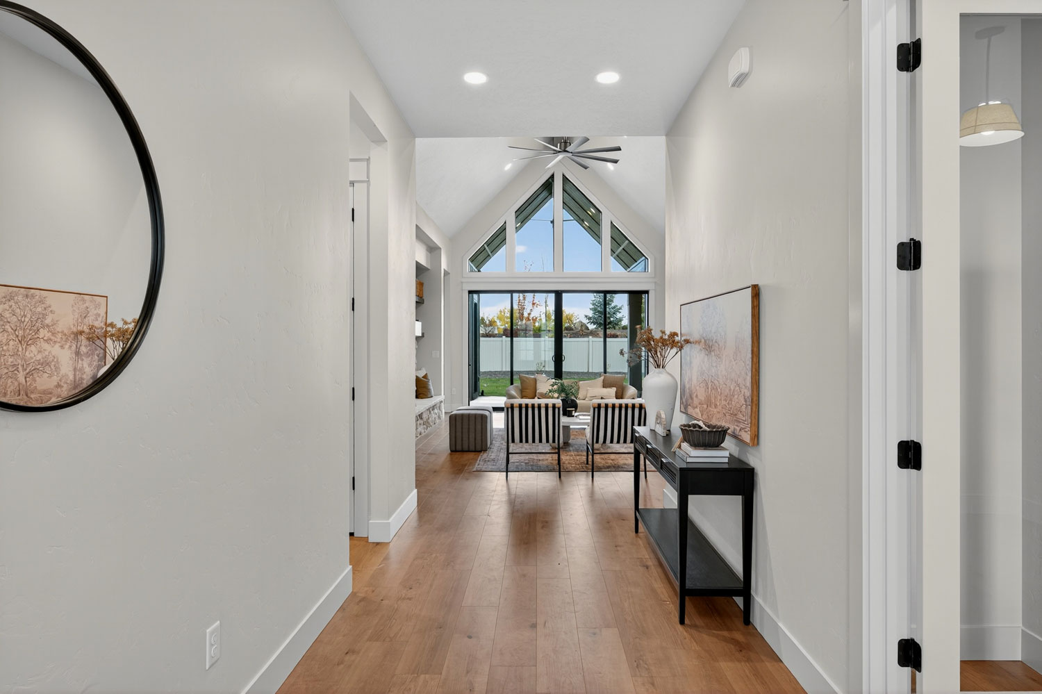 Bright entryway with warm hardwood floors, white walls, and round black-framed mirror leading into vaulted living room with floor-to-ceiling windows
