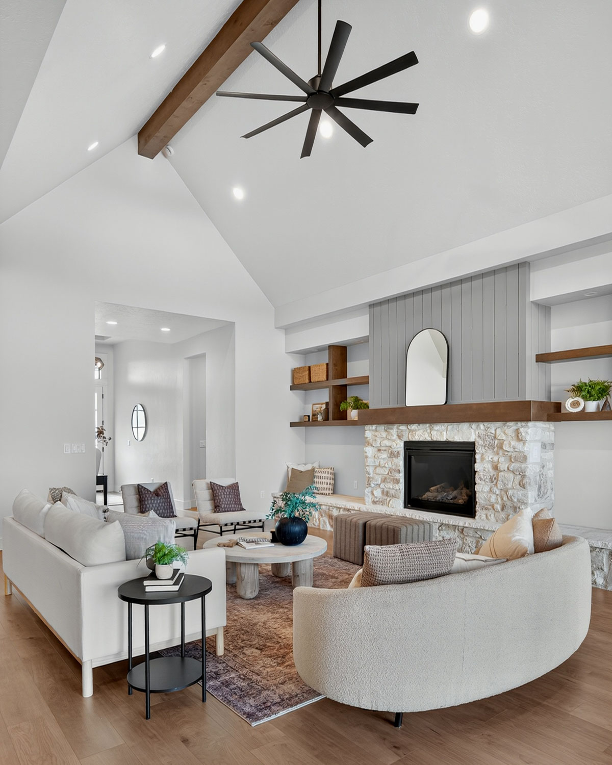 Vaulted ceiling living room with exposed wood beam, ceiling fan, stone fireplace, and curved boucle sofa on hardwood floors