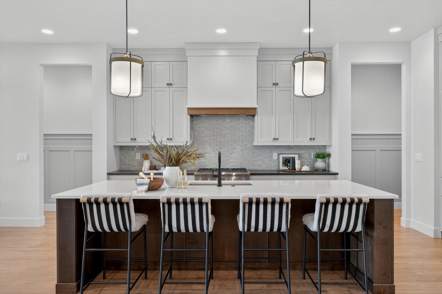 Kitchen island with four striped bar stools, white quartz countertop, dark wood base, pendant lantern lights, and shaker cabinetry