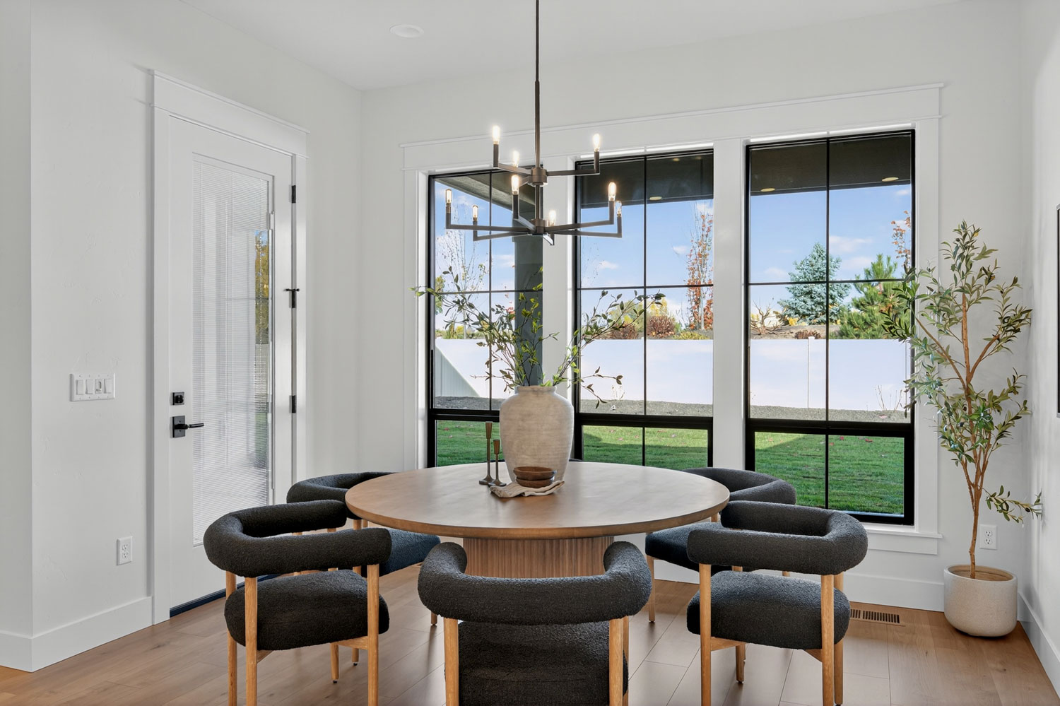 Breakfast nook with round wood dining table, dark boucle chairs, modern chandelier, and large windows overlooking the backyard