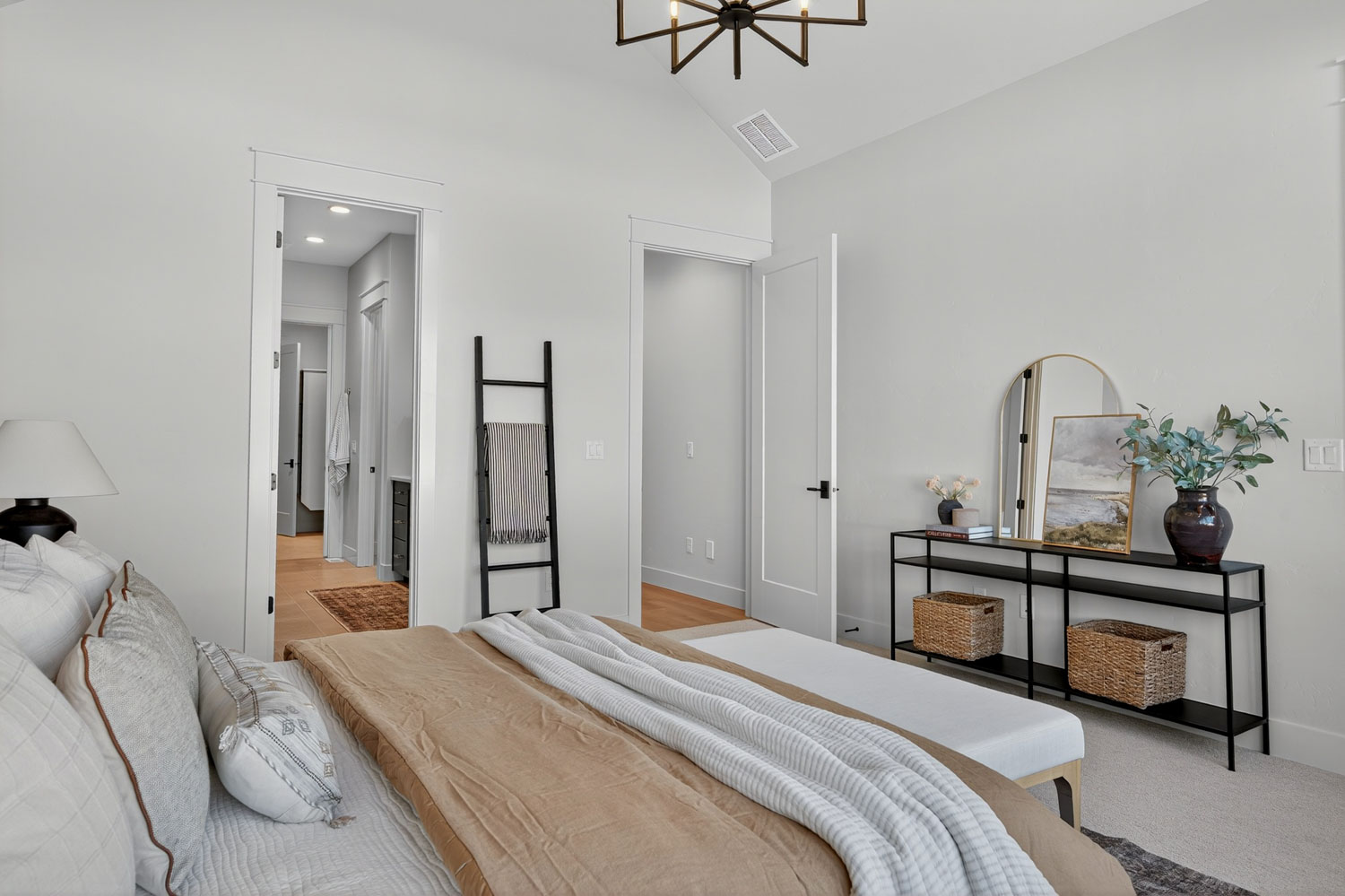 Master bedroom view with black metal console table, arched gold mirror, decorative blanket ladder, and doorway to en-suite bathroom