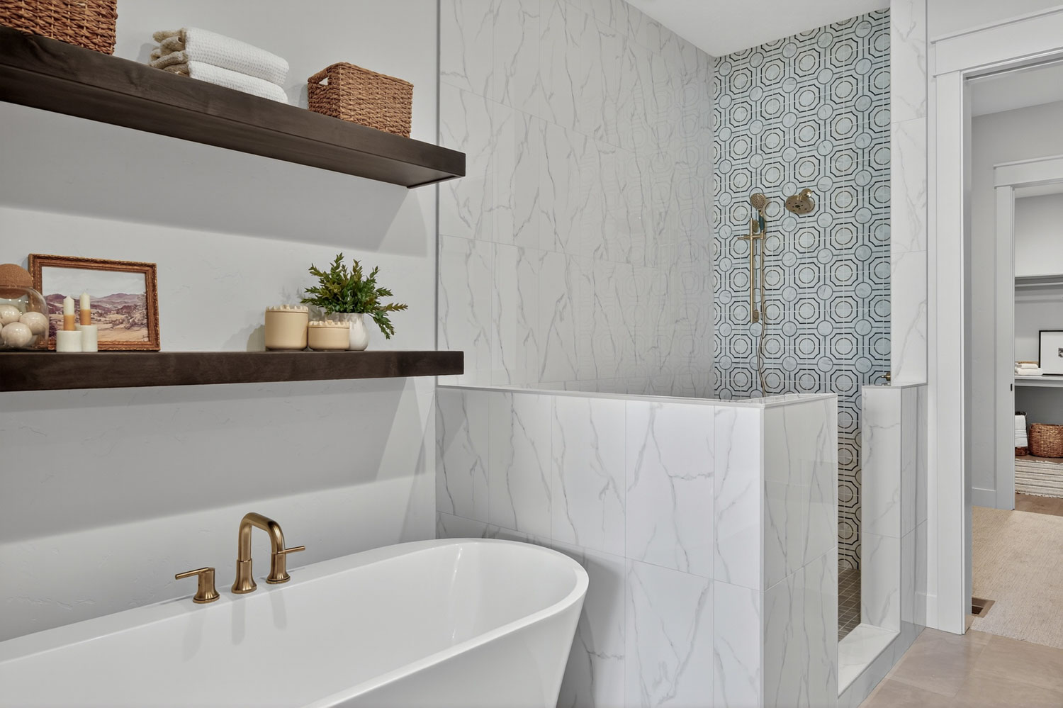 Freestanding white soaking tub with brushed gold faucet beneath dark wood floating shelves and marble-look wall tile