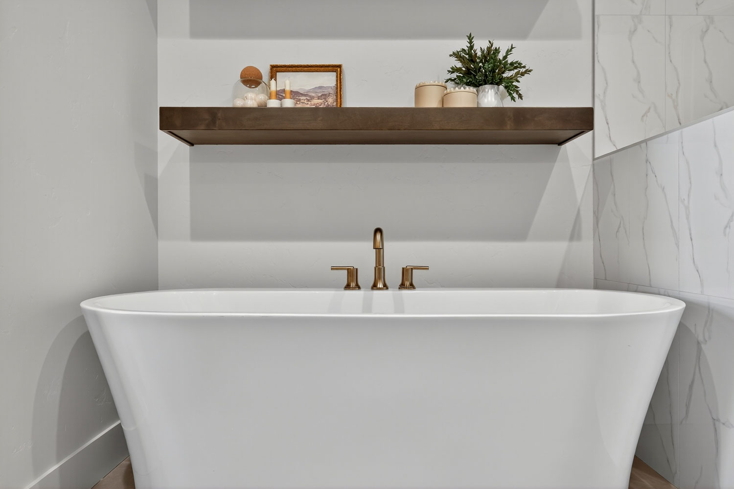 Close-up of freestanding bathtub with brushed gold fixtures, dark wood floating shelf, and marble-look porcelain wall surround