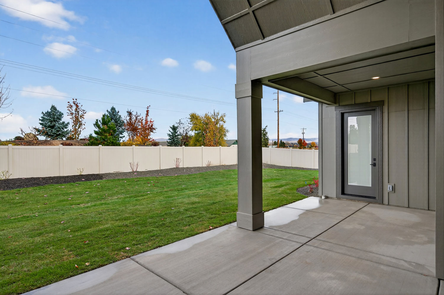 Covered back patio overlooking fenced backyard with green lawn, landscaped flower beds, and autumn-colored trees