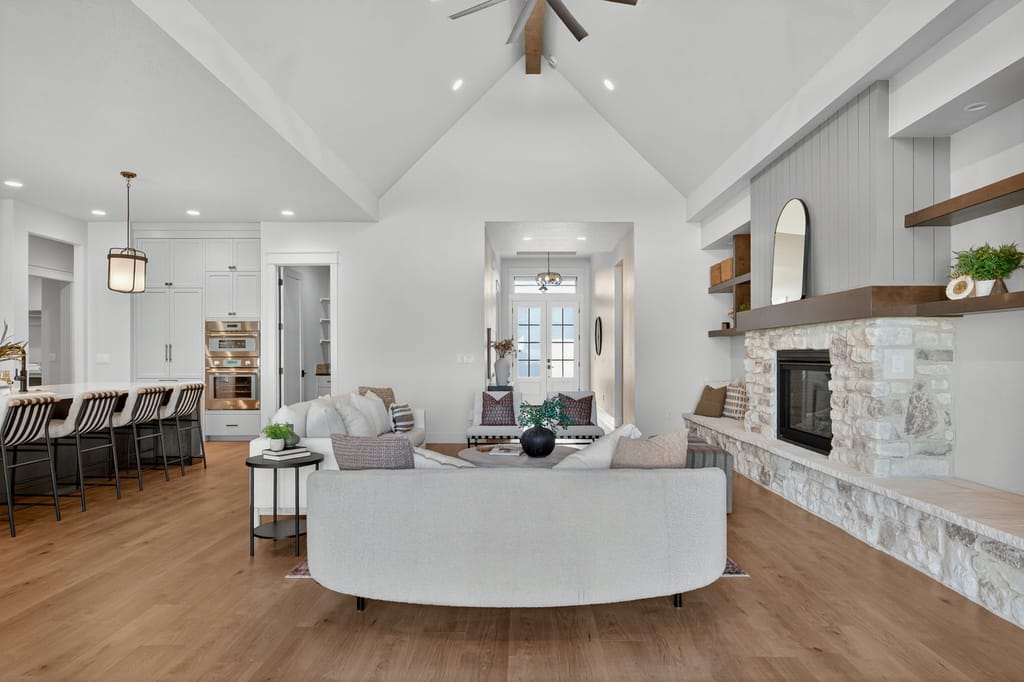Open-concept great room with vaulted ceilings featuring curved sofa, stone fireplace with floating wood shelves, and kitchen island with bar seating
