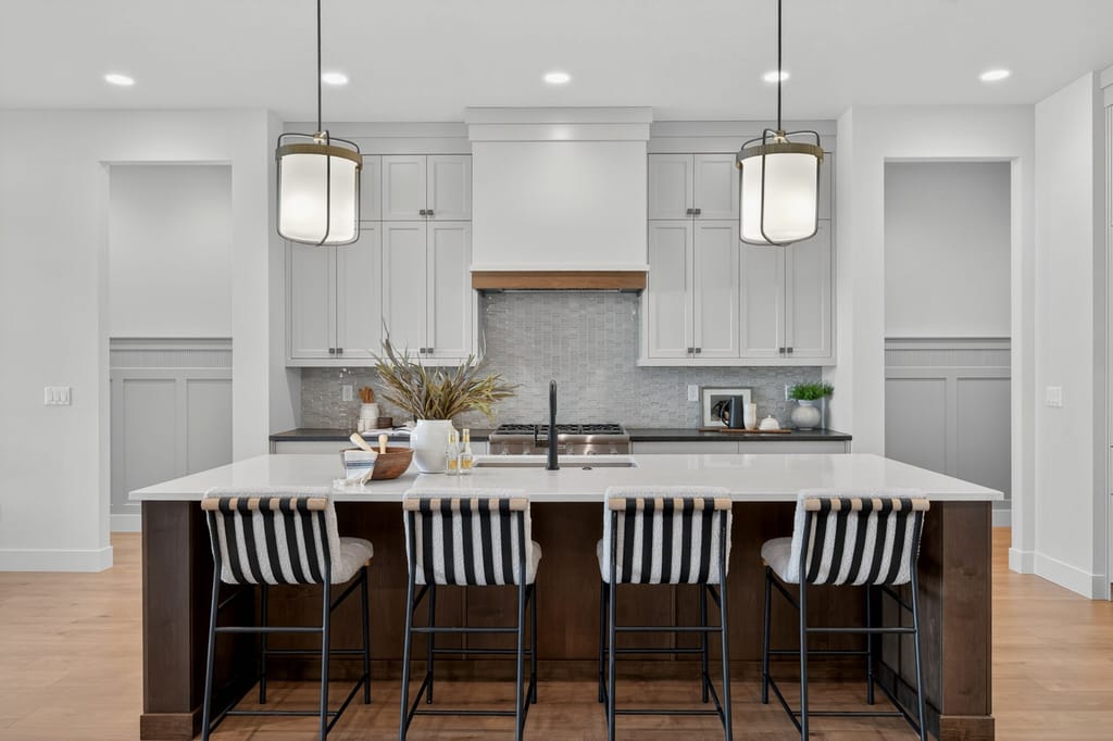 Kitchen island with four striped bar stools, white quartz countertop, dark wood base, pendant lantern lights, and shaker cabinetry