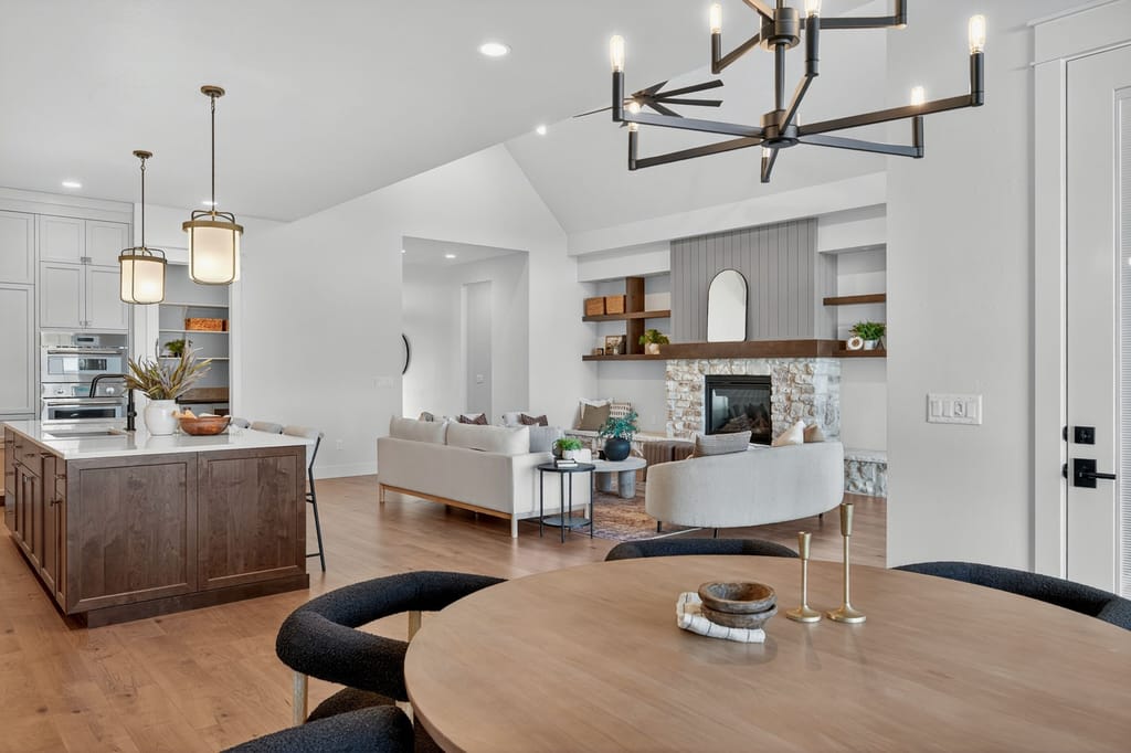 Open-concept dining and living area with modern chandelier, round wood table, kitchen island, stone fireplace, and vaulted ceilings