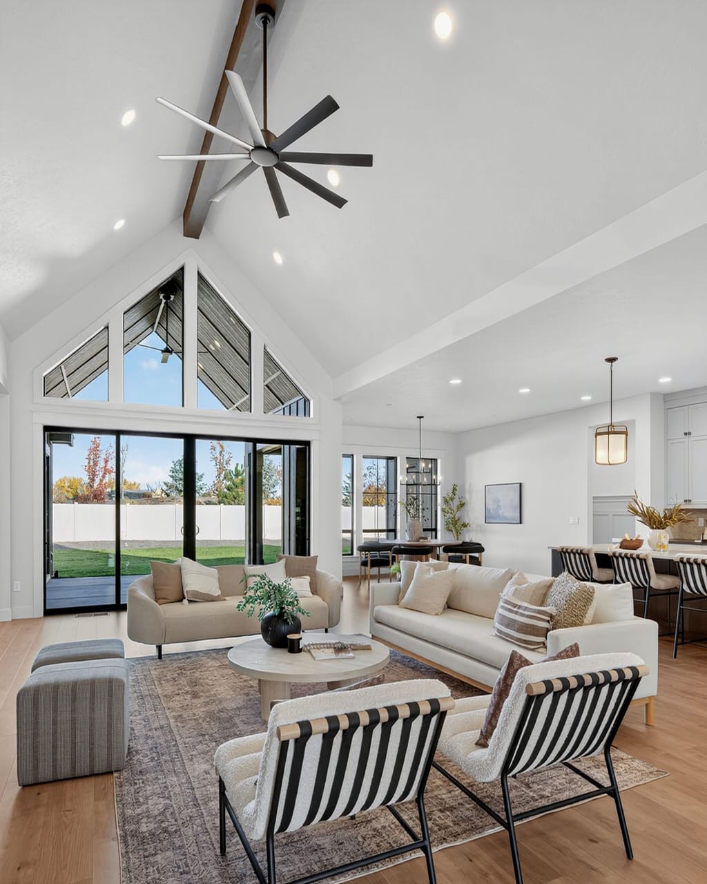 Expansive great room with vaulted ceiling, peaked glass wall, ceiling fan with wood beam, open kitchen, and neutral furnishings