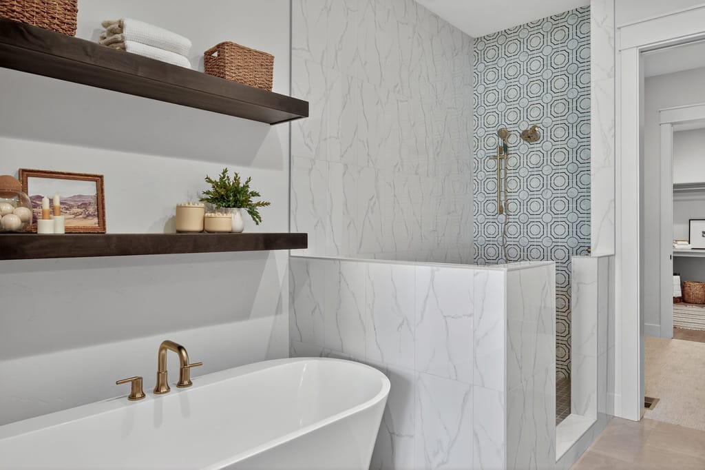 Freestanding white soaking tub with brushed gold faucet beneath dark wood floating shelves and marble-look wall tile