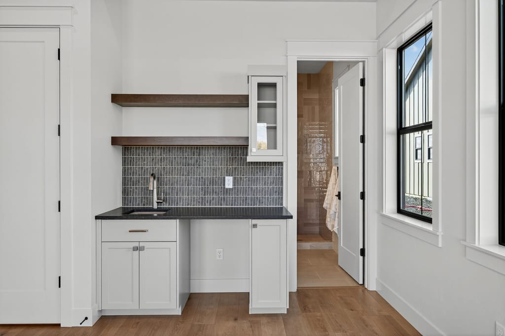 Built-in wet bar with white shaker cabinets, dark granite countertop, green stacked tile backsplash, and floating wood shelves